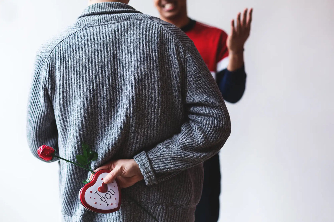 Person in a gray knit sweater holding a heart decoration behind their back.